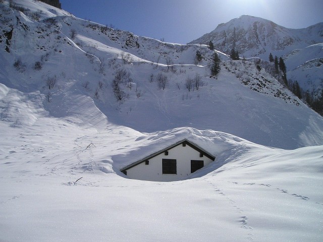 A house tucked deep into the snow in a mountain valley, the sun shines above in this photo