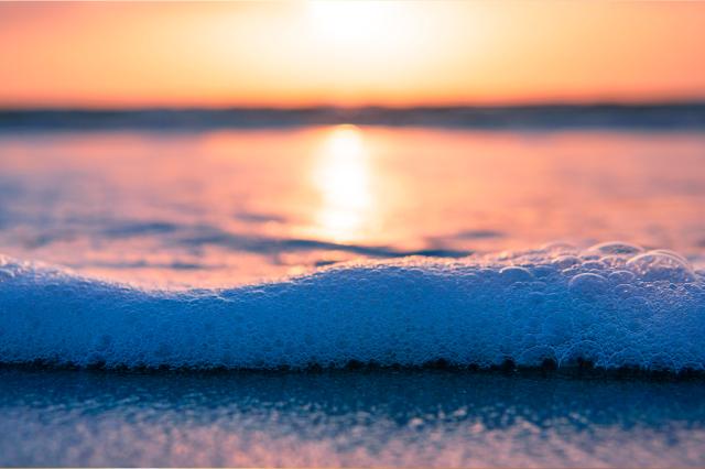 photo of a wave reaching the shore on a body of water where the sun is low on the horizon, nearly sunset, and there are bubbles in the water